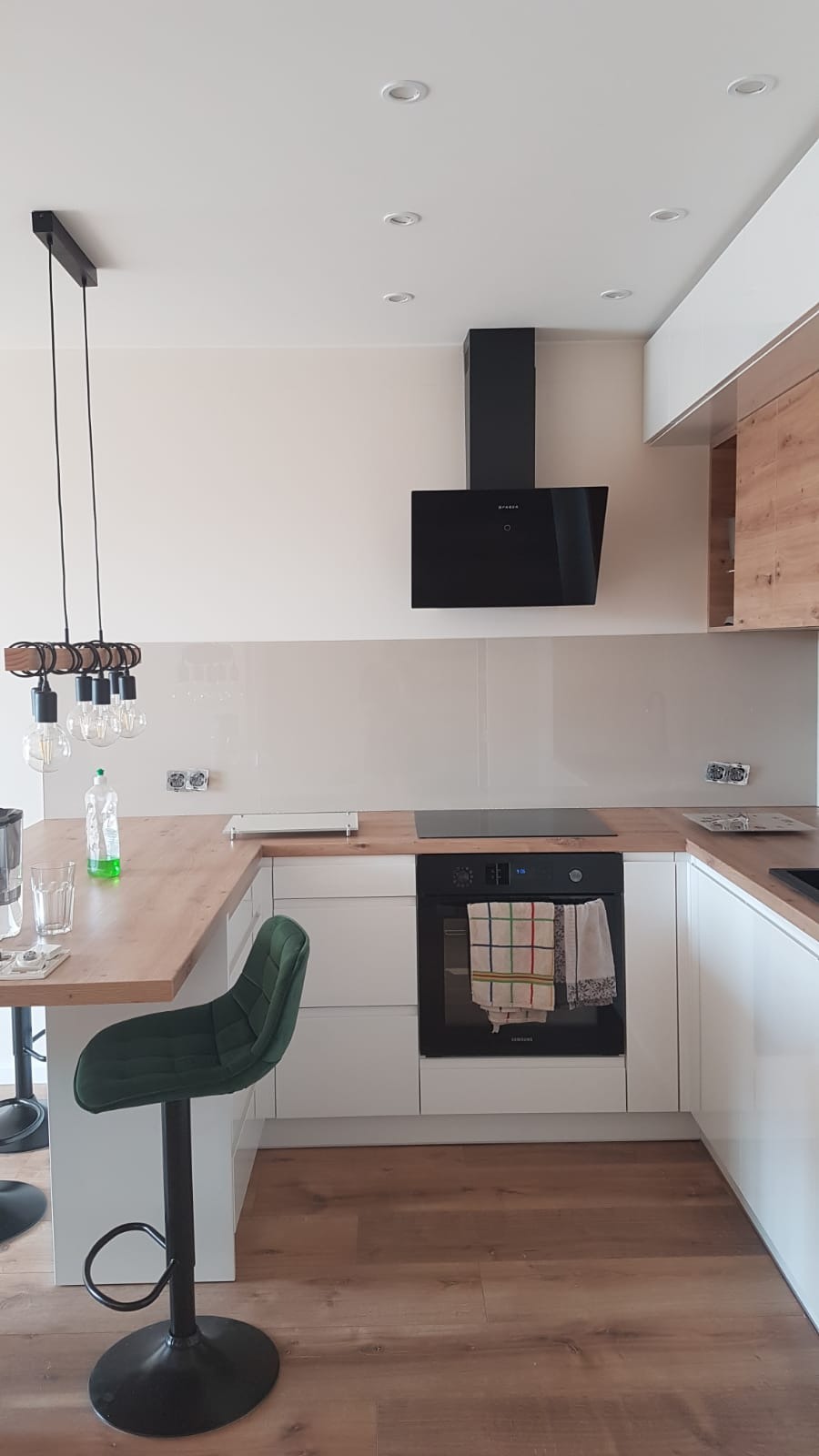 Nowoczesna kuchnia z białymi szafkami, drewnianym blatem i czarnym okapem, oświetlona wiszącą lampą z żarówkami Edisona i zielonym krzesłem barowym.