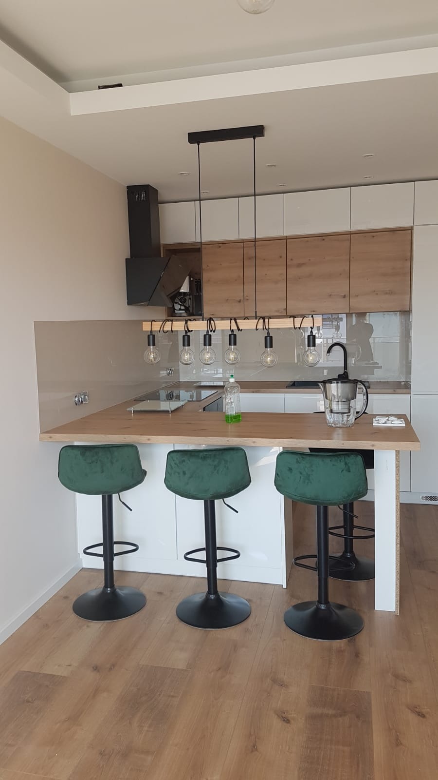 Nowoczesna kuchnia z wyspą, drewnianymi szafkami i białymi frontami, oświetlona lampą z żarówkami Edisona i wyposażona w zielone hokery.