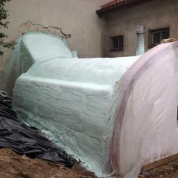 Techniczne Usługi Natryskowe - Ocieplanie pianką PUR: fragment ściany budynku po natrysku pianki, widoczne zabezpieczenie folią. Detal: nierównomierna struktura pianki, czarna folia ochronna na ziemi.