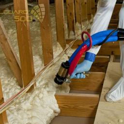 Techniczne Usługi Natryskowe - Aplikacja pianki PUR w przestrzeni poddasza. Widoczne drewniane elementy konstrukcyjne i osoba w kombinezonie ochronnym, aplikująca pianę za pomocą specjalistycznego sprzętu.