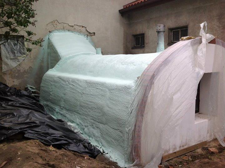 Ocieplanie pianką PUR: fragment fundamentu budynku pokryty jasną pianką. Widoczne czarne folie ochronne i fragment ściany z oknami w tle.