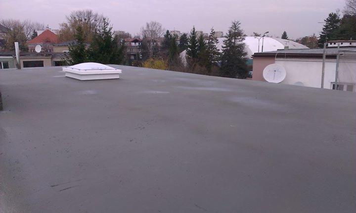 Szary dach budynku z świetlikiem dachowym na tle zabudowy miejskiej i pochmurnego nieba. Widoczny budynek z anteną satelitarną.