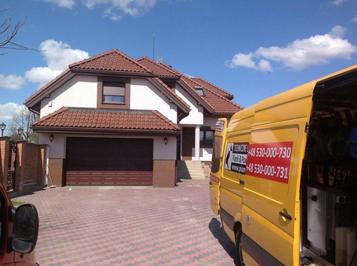 Dwukondygnacyjny dom z garażem i żółty samochód serwisowy z numerem telefonu na tle błękitnego nieba z chmurami.