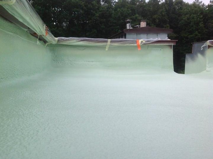 Świeżo nałożona, jasna pianka PUR na płaskiej powierzchni dachu, zabezpieczona folią i taśmą malarską przy krawędziach. Widoczne drzewa w tle.