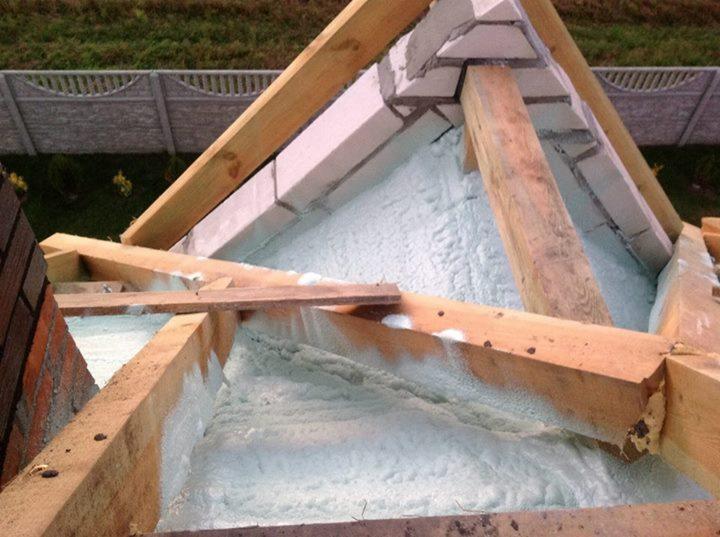 Ocieplenie poddasza pianką PUR: biała izolacja między drewnianymi krokwiami i ceglanym kominem, widok z góry na ocieplony dach z elementami konstrukcyjnymi.