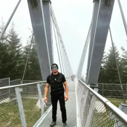 Mężczyzna w czarnym ubraniu i okularach przeciwsłonecznych pozuje na metalowym moście wiszącym z siatkowymi barierkami, w tle zalesiony krajobraz w mglisty dzień.