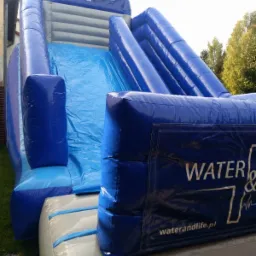 Niebiesko-szara dmuchana zjeżdżalnia z logo 'WATER & LIFE' ustawiona na trawie, widok z dołu, w tle budynek i drzewa.