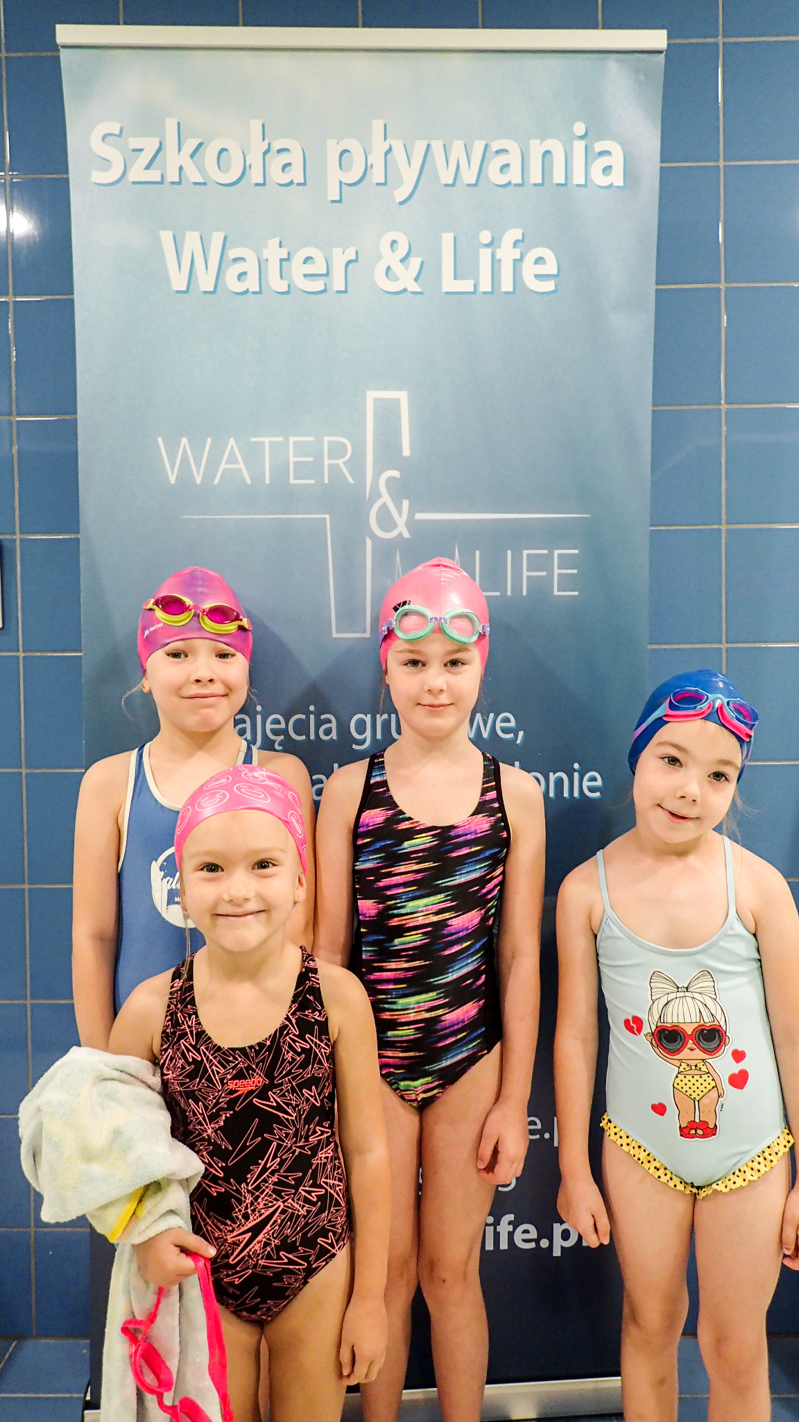 Grupa czterech dziewczynek w strojach kąpielowych i czepkach, pozujących przed banerem Szkoły Pływania Water & Life na tle niebieskich płytek basenowych.
