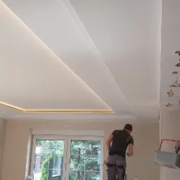 Pracownik na drabinie podczas malowania sufitu z podświetleniem LED w wyremontowanym pokoju, widoczne narzędzia malarskie i grzejnik pod oknem.