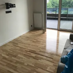 Jasny parkiet ułożony w jodełkę francuską w pokoju z wyjściem na balkon, widoczne gniazdka i uchwyt na telewizor na ścianie.