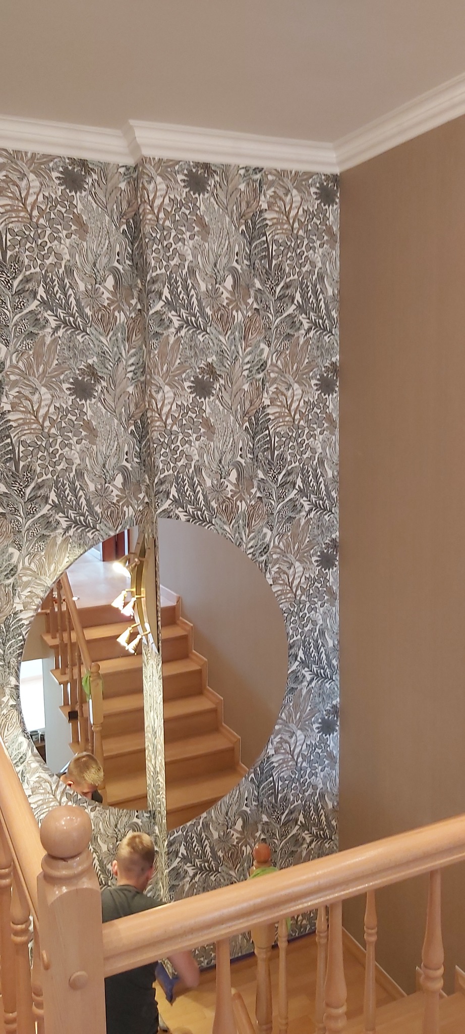Wzorzysta tapeta w odcieniach szarości i brązu na ścianie przy schodach, częściowo widoczna w okrągłym lustrze, pracownik wygładza tapetę na dole ściany.