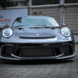 Błyszczący, czarny Porsche 911 z włączonymi światłami dziennymi, zaparkowany przed budynkiem z szyldem 'autoklinika', prezentujący detale karoserii i tuning.