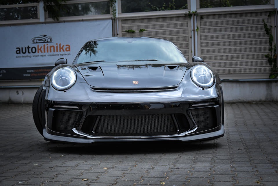 Błyszczący, czarny Porsche 911 z włączonymi światłami dziennymi, zaparkowany przed budynkiem z szyldem 'autoklinika', prezentujący detale karoserii i tuning.