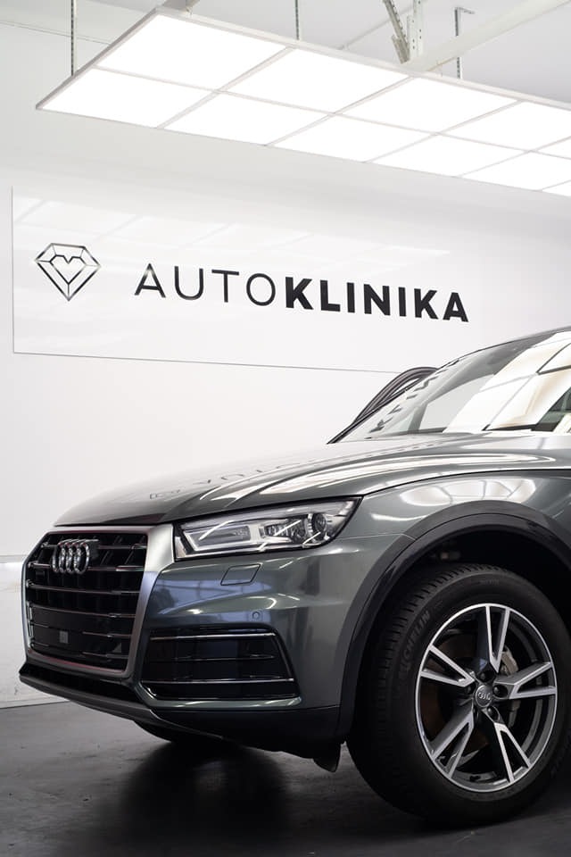 Szary Audi Q5 na tle białej ściany z logo Autoklinika, widoczne przednie światło, grill i opona Michelin.
