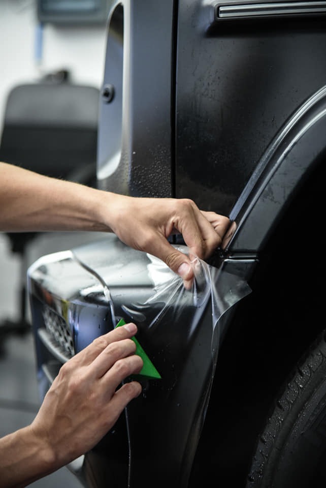Aplikacja folii ochronnej na czarny, matowy samochód terenowy; widoczne dłonie pracownika i zielony aplikator.