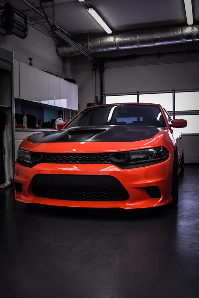 Pomarańczowy samochód z czarną maską w garażu z białymi szafkami w tle, widok z przodu, oświetlenie górne.