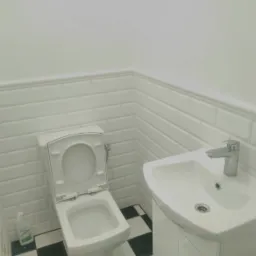 Biała toaleta z otwartą klapą i umywalka w małej łazience wykończonej białymi płytkami typu metro i czarno-białą szachownicą na podłodze. Świeżo pomalowane ściany.