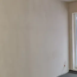 Świeżo położona wylewka podłogowa w szarym kolorze, ściany otynkowane, widoczne niedoskonałości na suficie, wystające kable elektryczne ze ściany.