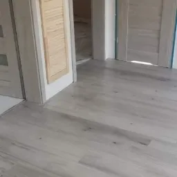 Jasnoszara podłoga z paneli laminowanych, widoczne dwie pary drzwi wewnętrznych, jedne z drewnianymi żaluzjami.