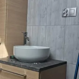 Nowoczesna łazienka w Kościanie z okrągłą umywalką nablatową, armaturą i szafką z szufladami w odcieniach drewna, tło stanowią szare płytki imitujące drewno.