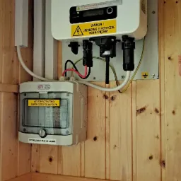 Biały falownik Sofar Solar zamontowany na drewnianej ścianie z zabezpieczeniami elektrycznymi ETI, ostrzeżenie o napięciu i naklejka 'Fotowoltaika! Prąd za darmo!'