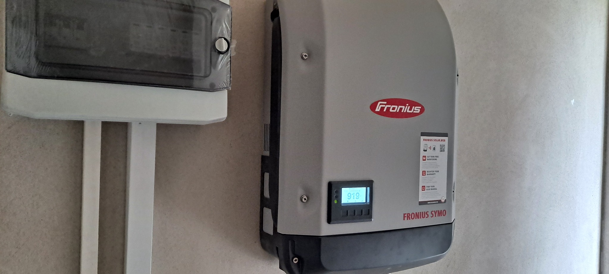 Szary inwerter Fronius Symo zamontowany na ścianie obok skrzynki elektrycznej z widocznym wyświetlaczem wskazującym wartość 919.