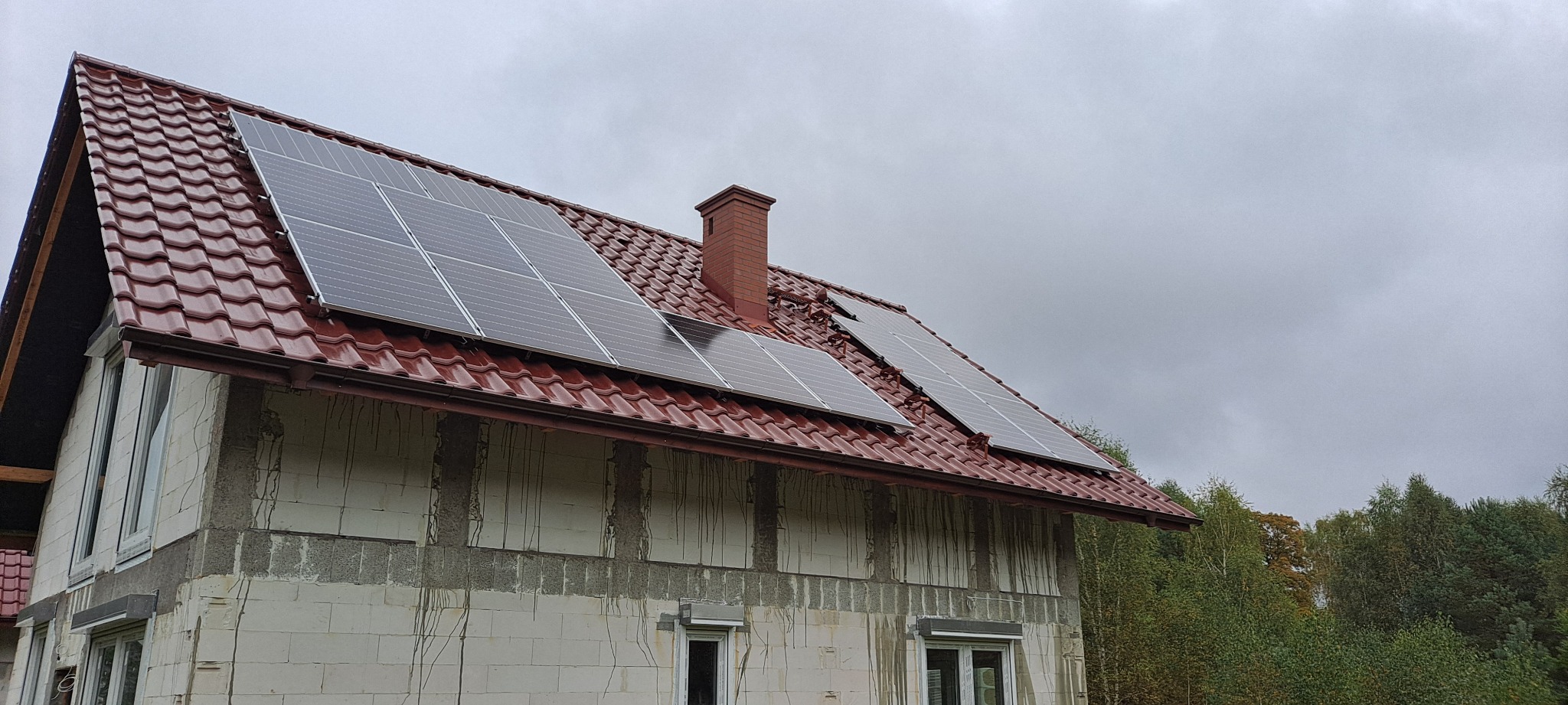 Dom w trakcie budowy z zamontowanymi panelami słonecznymi na czerwonym dachu, widok z dołu na pochmurne niebo.