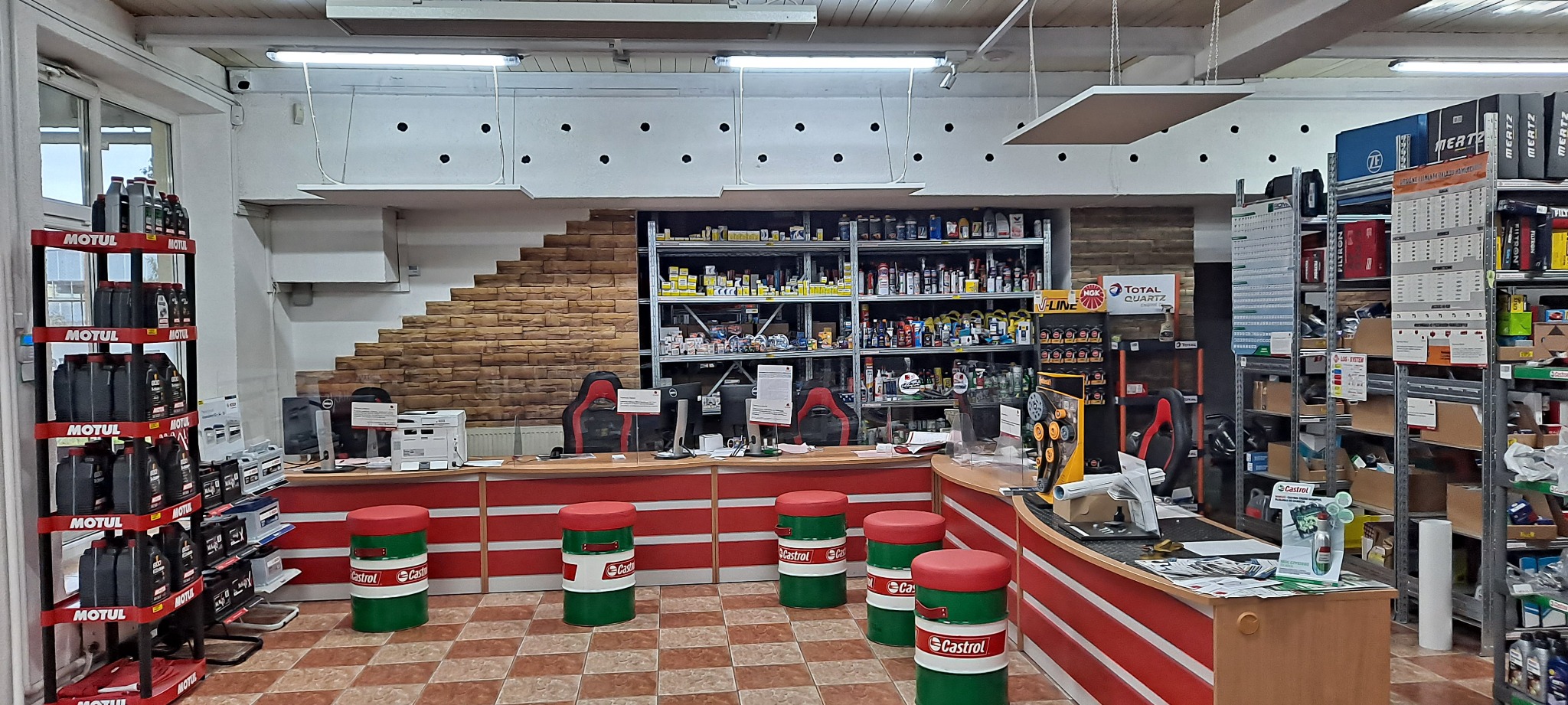 Wnętrze sklepu z częściami samochodowymi i olejami, widoczne regały z produktami Motul i Castrol, stanowisko obsługi klienta z krzesłami w kształcie beczek Castrol.