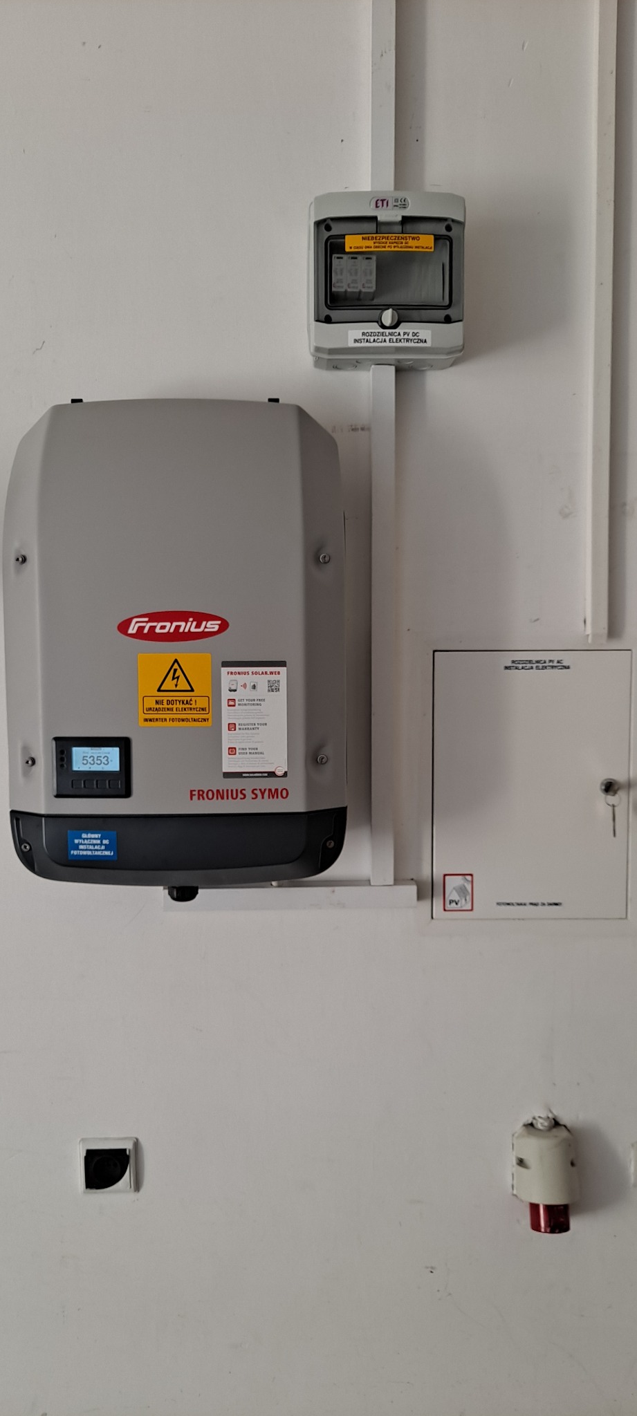 Szary inwerter Fronius Symo zamontowany na białej ścianie obok skrzynki rozdzielczej PV DC i licznika energii elektrycznej. Widoczne ostrzeżenie o napięciu i naklejka Fronius Solar.web.