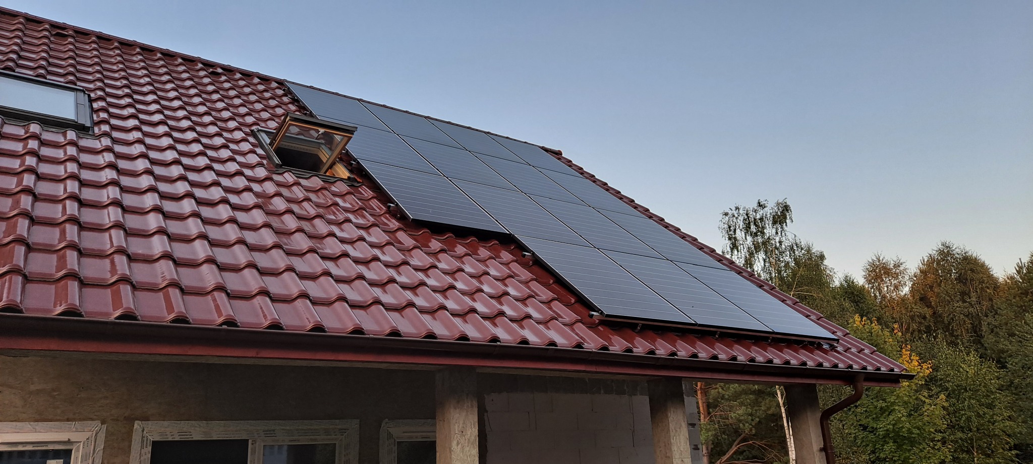Panele słoneczne na dachu domu z czerwoną dachówką, widoczne otwarte okno dachowe i fragment lasu w tle.