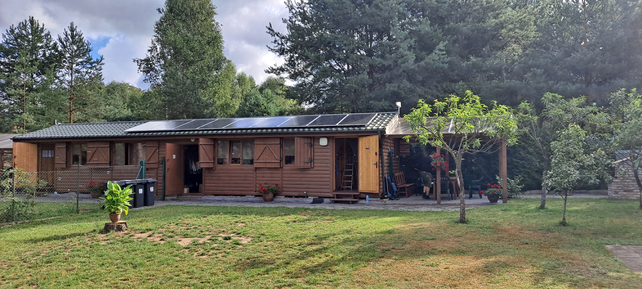 Brązowy domek letniskowy z panelami słonecznymi na dachu, otoczony zielonym trawnikiem i drzewami.