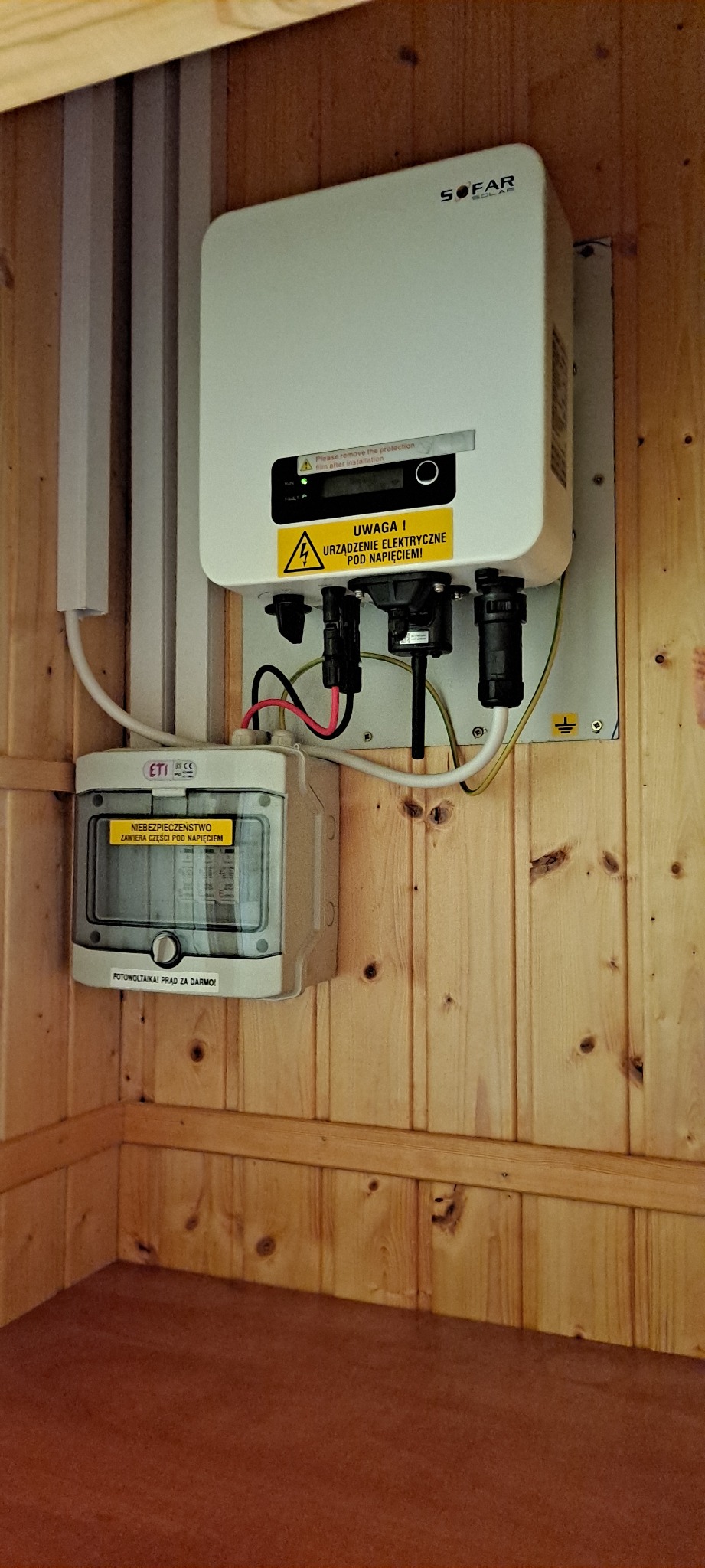 Biały falownik Sofar Solar zamontowany na drewnianej ścianie z zabezpieczeniami elektrycznymi ETI, ostrzeżenie o napięciu i naklejka 'Fotowoltaika! Prąd za darmo!'