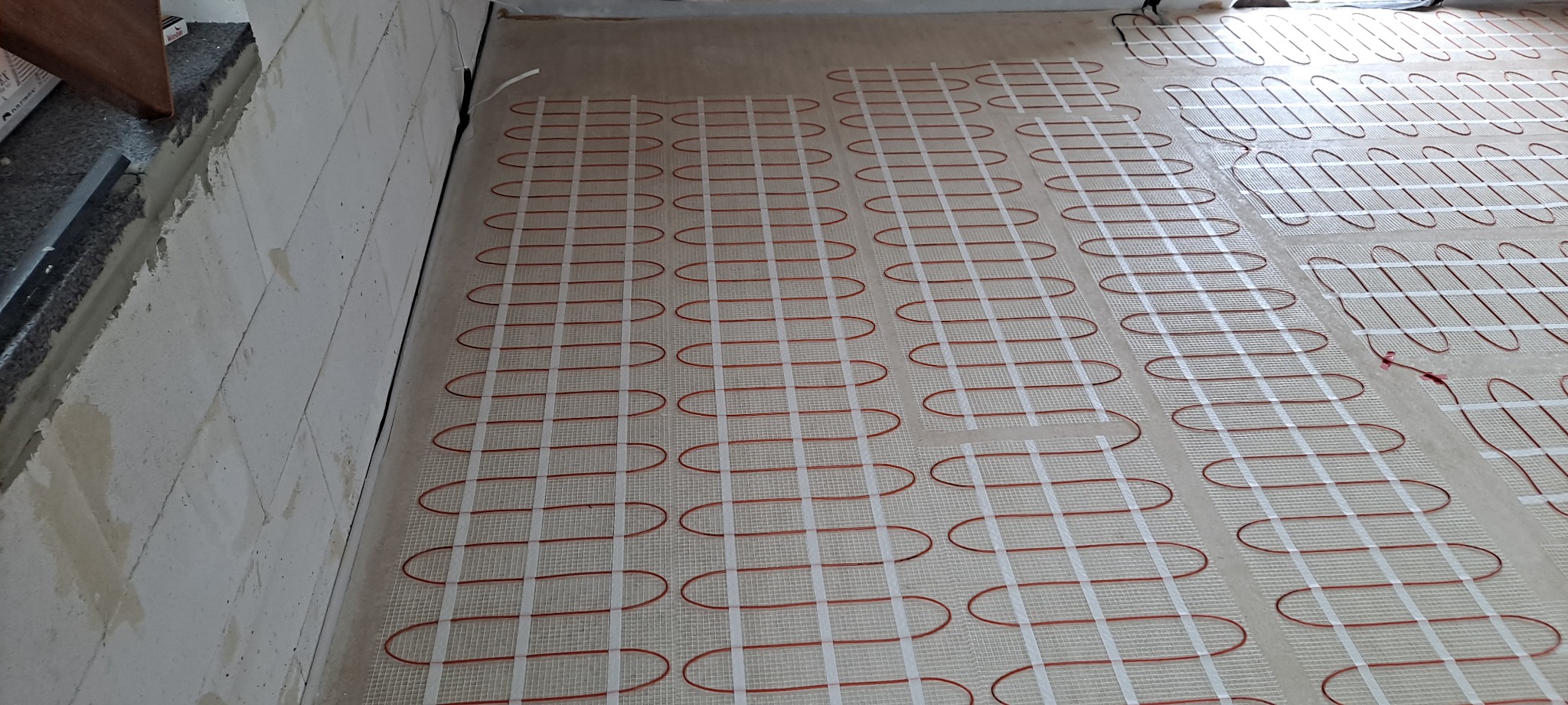 Rozłożona mata grzewcza z czerwonymi przewodami i białymi taśmami mocującymi na podłożu, przygotowana do wylewki.