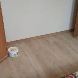 Wnętrze pokoju z białą ścianą po świeżym pomalowaniu, widoczna puszka farby przy listwie przypodłogowej i fragment wzorzystej tkaniny na podłodze.