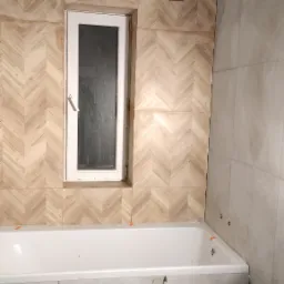 Łazienka w trakcie wykańczania: ściana z płytek ułożonych w jodełkę wokół okna, częściowo obłożona wanna z widocznymi klipsami do płytek, surowy sufit z widocznymi otworami na oświetlenie.