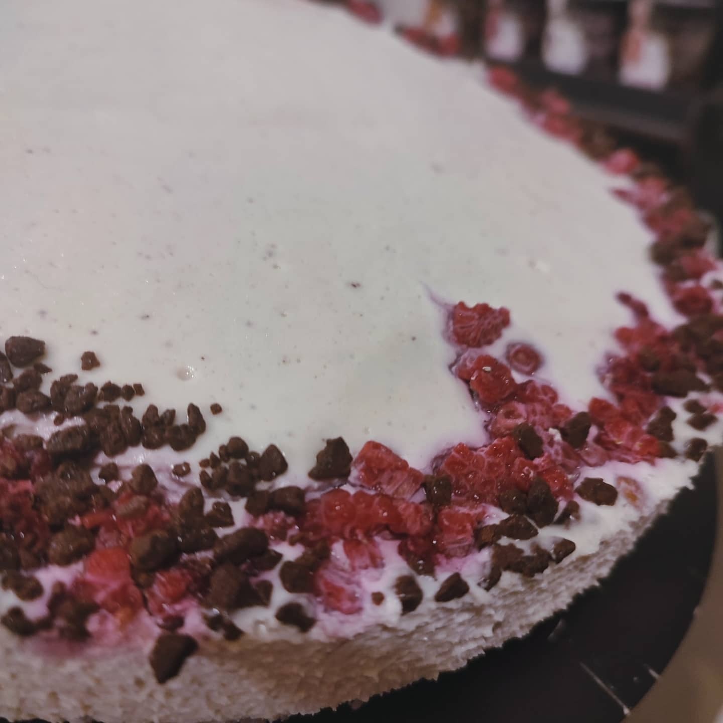 Tort z białą polewą, malinami i czekoladowymi kruszonkami, widok z bliska, na czarnym talerzu.