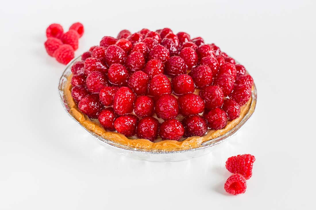 Tarta z malinami w srebrnej formie, błyszcząca glazura, kilka malin obok na białym tle.