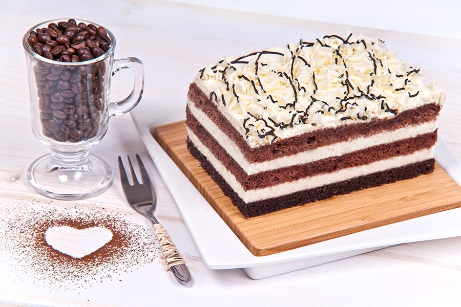 Kawałki warstwowego tortu czekoladowego z kremem i kawałkami białej czekolady na wierzchu, podane na drewnianej desce, obok filiżanka z ziarnami kawy i widelec z owiniętą rączką, a także serce...