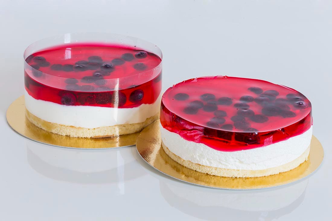 Dwa okrągłe ciasta warstwowe z biszkoptem, kremem, czerwoną galaretką i borówkami na złotych podkładkach, jedno w przezroczystej obręczy.