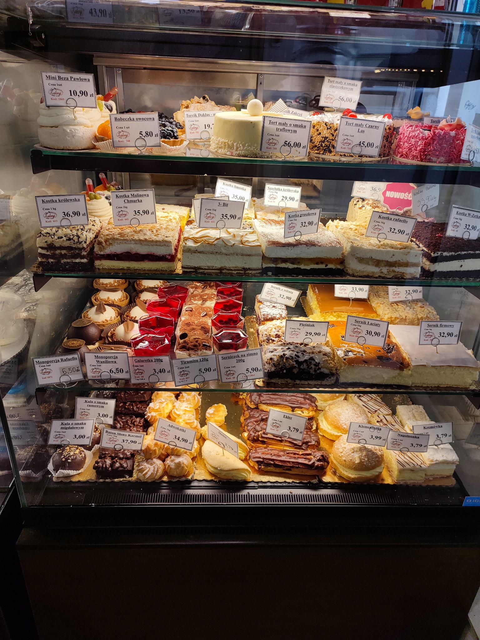Szeroki kadr witryny cukierniczej z różnorodnymi ciastami, tortami i deserami, każdy z etykietką cenową, oświetlonymi ciepłym światłem.
