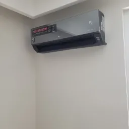 Szary klimatyzator LG Dual Inverter zamontowany na jasnej ścianie, widoczny z boku w rogu pomieszczenia, z naklejką informującą o oszczędności energii.