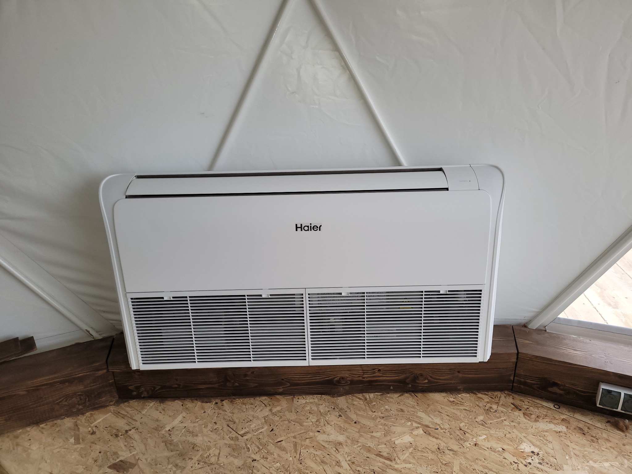 Biała jednostka wewnętrzna klimatyzatora marki Haier zamontowana w pomieszczeniu o konstrukcji namiotowej, z widocznym drewnianym obramowaniem i podłogą z płyty OSB.