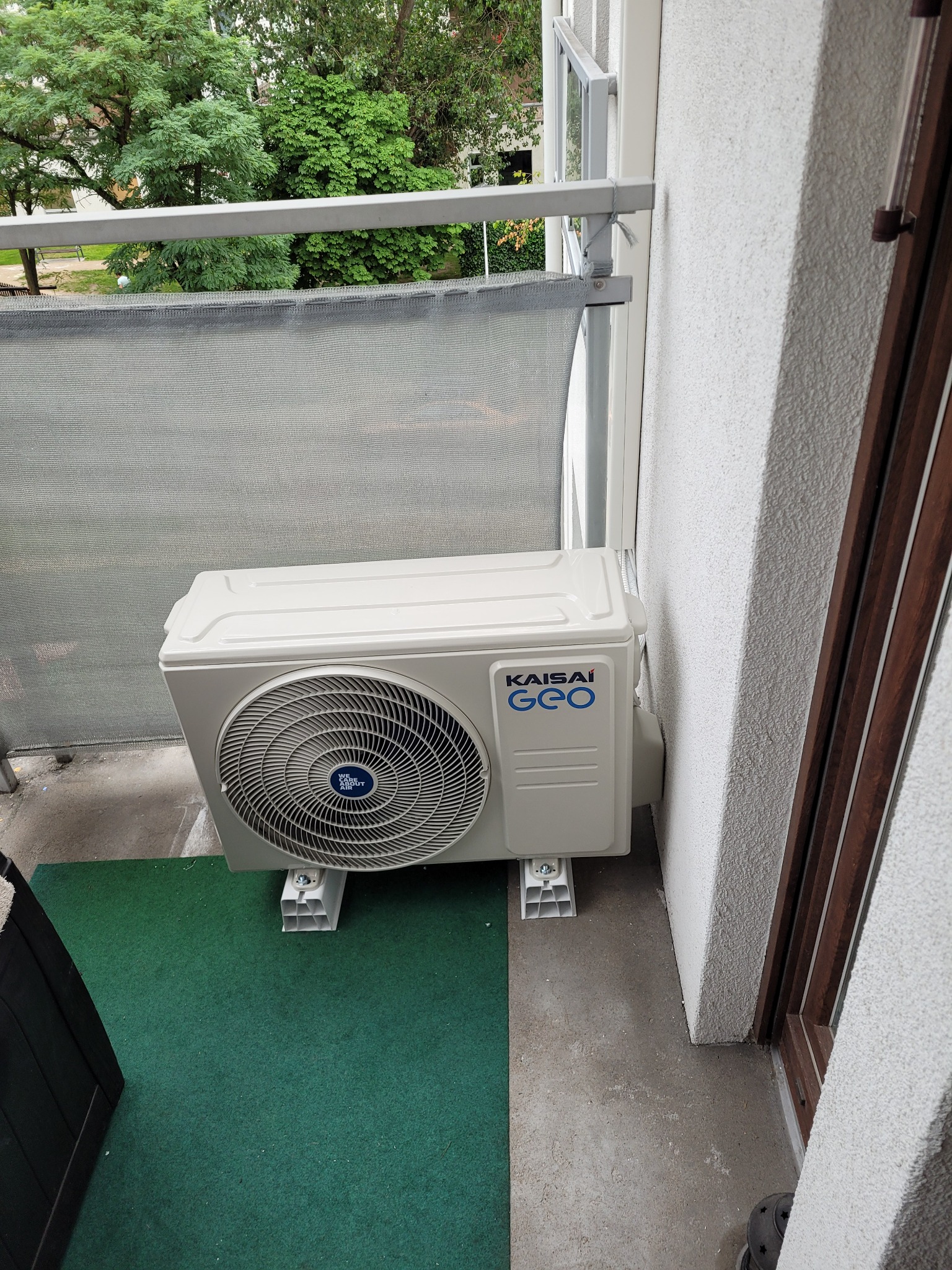 Zewnętrzna jednostka klimatyzatora Kaisai GEO zamontowana na balkonie, widoczne wsporniki montażowe i fragment balustrady z siatką zacieniającą.