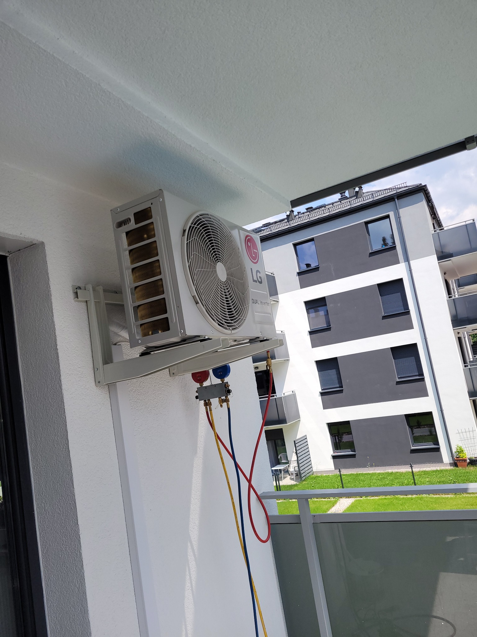 Biała jednostka zewnętrzna klimatyzatora LG Dual Inverter zamontowana na balkonie nowoczesnego budynku, podłączona do manometrów serwisowych z kolorowymi przewodami.