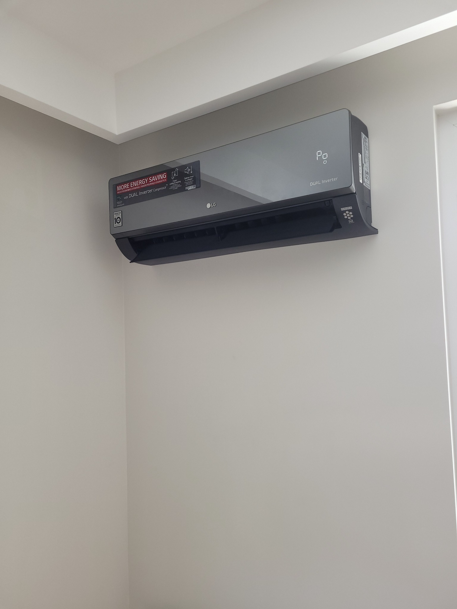 Szary klimatyzator LG Dual Inverter zamontowany na jasnej ścianie, widoczny z boku w rogu pomieszczenia, z naklejką informującą o oszczędności energii.
