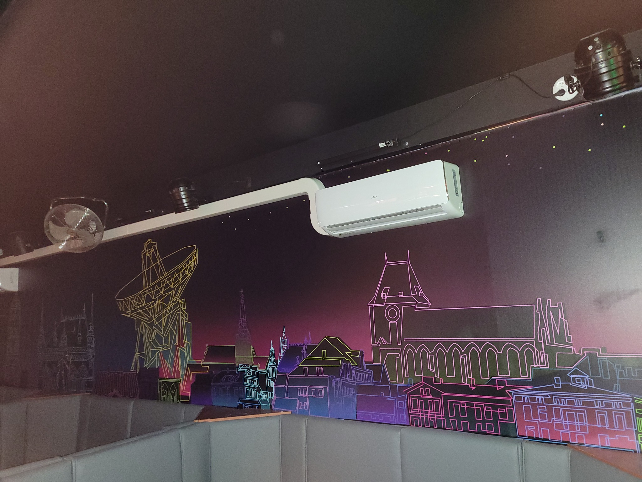 Biała jednostka klimatyzacyjna AUX zamontowana na ścianie z neonowym rysunkiem panoramy miasta i anteny satelitarnej, oświetlenie punktowe na suficie.