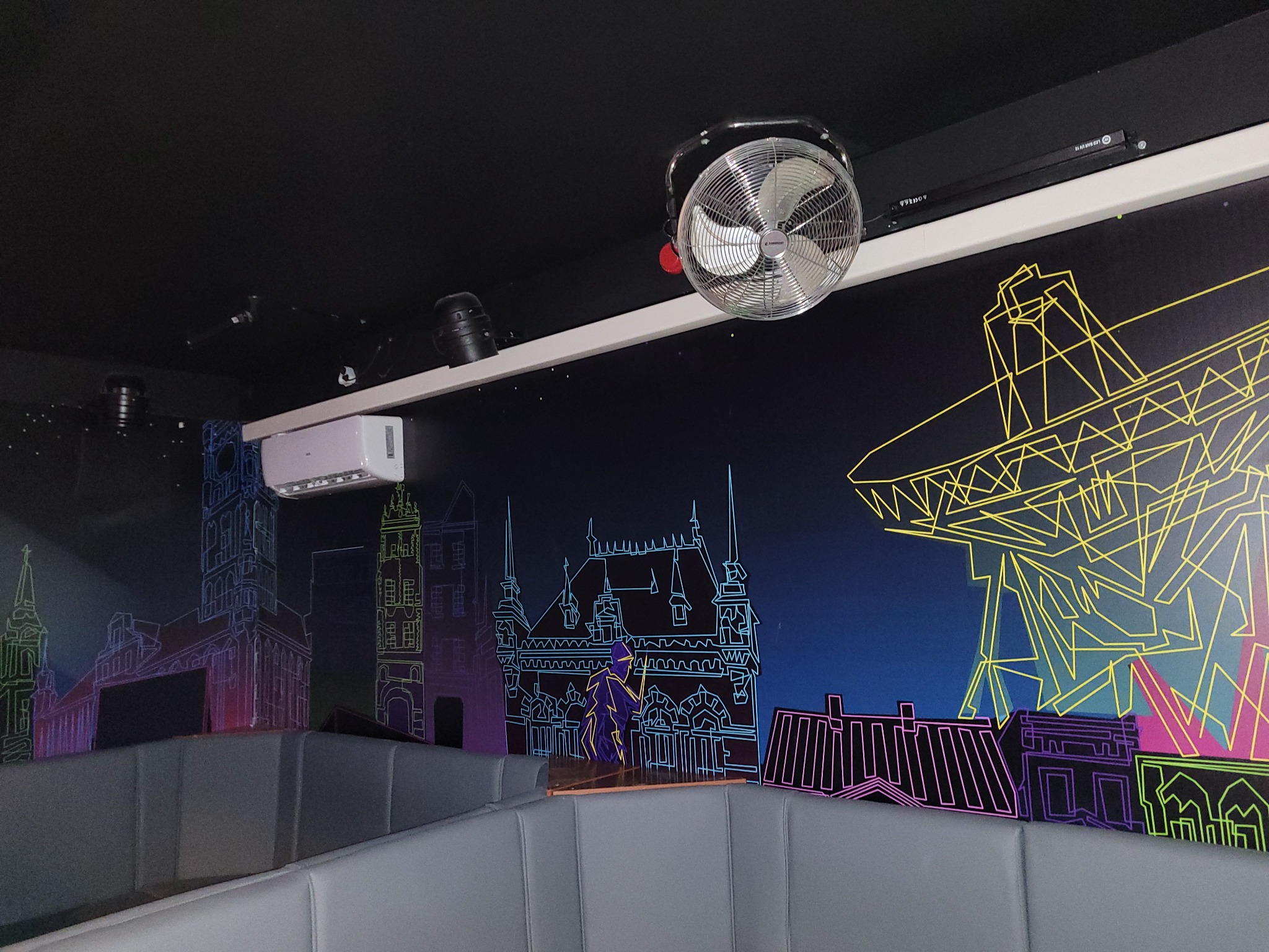 Wnętrze lokalu z muralem przedstawiającym architekturę miejską, wiszący wentylator sufitowy i klimatyzator ścienny.