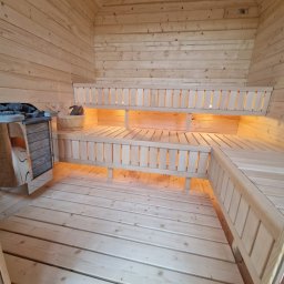 GDZborowski - Wnętrze nowej sauny z jasnego drewna, z dwupoziomowymi ławami i ciepłym, ukrytym oświetleniem LED pod nimi. Widoczny piec z kamieniami, wiaderko i chochla.