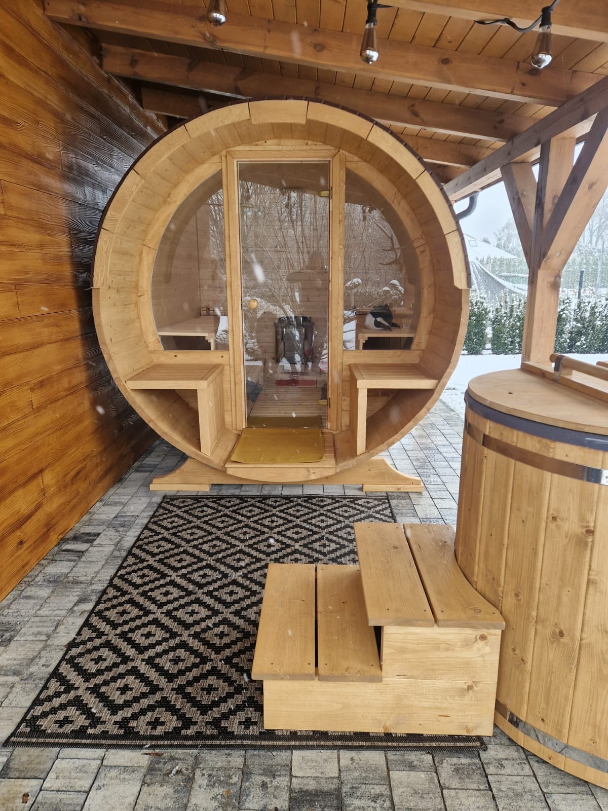 Drewniana sauna w kształcie beczki z przeszklonymi drzwiami, obok drewniana balia. Na zewnątrz pada śnieg, widoczny wzorzysty dywanik i drewniane schodki.