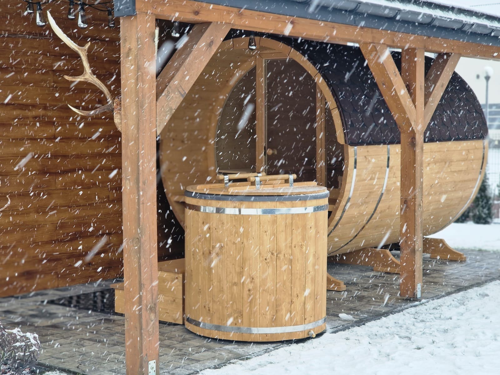Drewniana sauna beczka i balia w zimowej scenerii, padający śnieg. Sauna zadaszona, obok balii widoczne uchwyty pokrywy. Na ścianie sauny poroże jelenia.